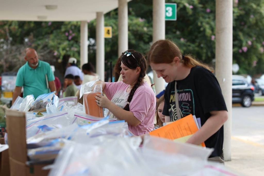 Two school staff members and a parent or volunteer interacting outdoors under a covered walkway near numbered posts and green trees.