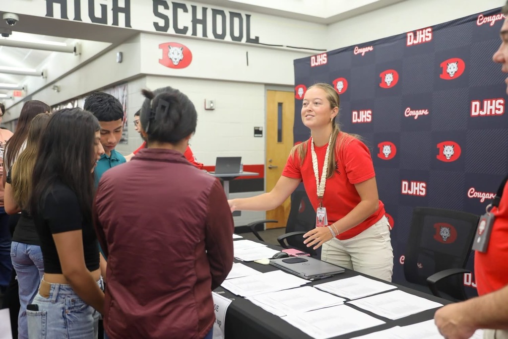A smiling school staff member wearing a red lanyard stands at an indoor registration or check-in table in front of a DJHS backdrop.