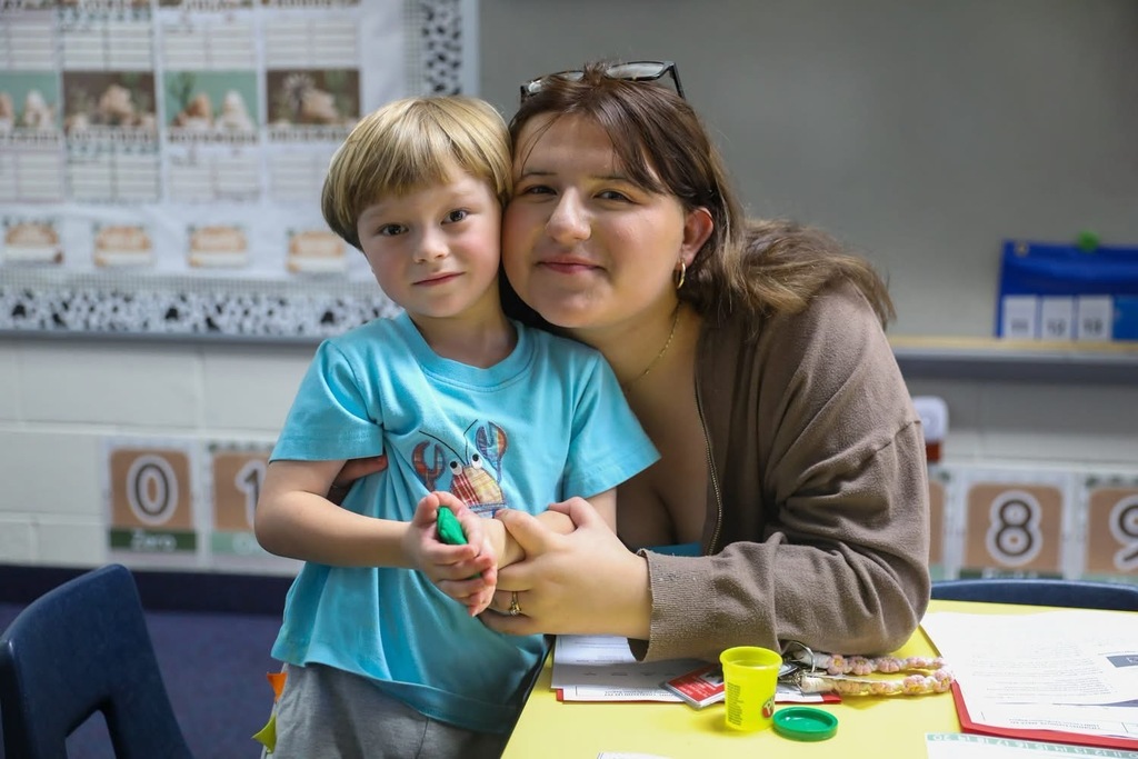 A young smiling boy in a light blue t-shirt standing beside a teenage girl sitting at a classroom table with playdough, small plastic cups, and learning materials.