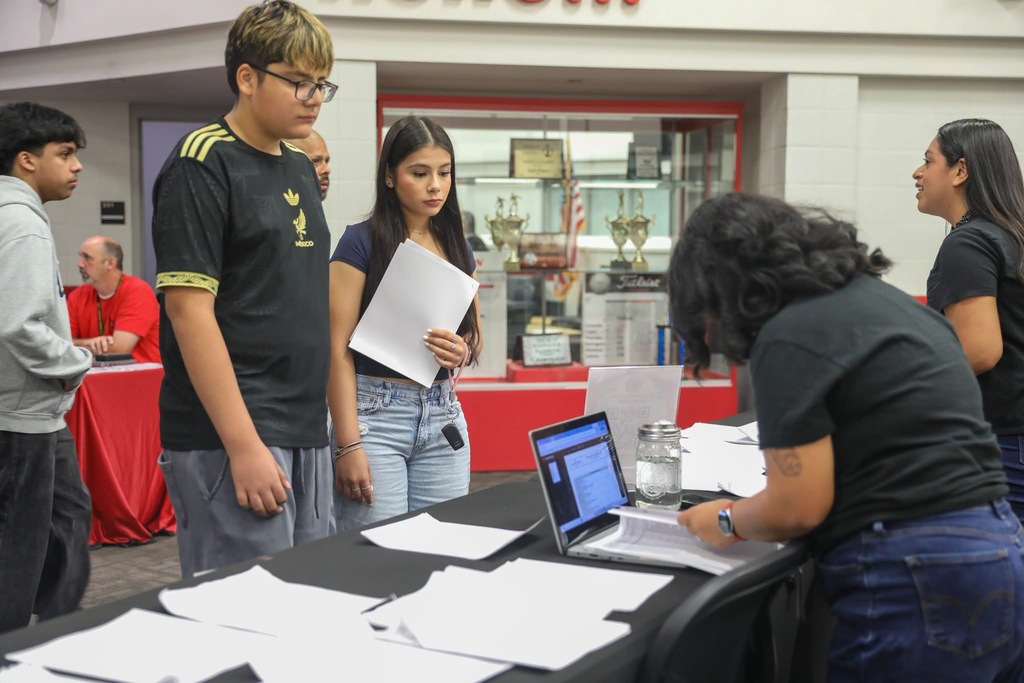 A young man wearing glasses and a dark soccer jersey stands with students and staff in a brightly lit school lobby.