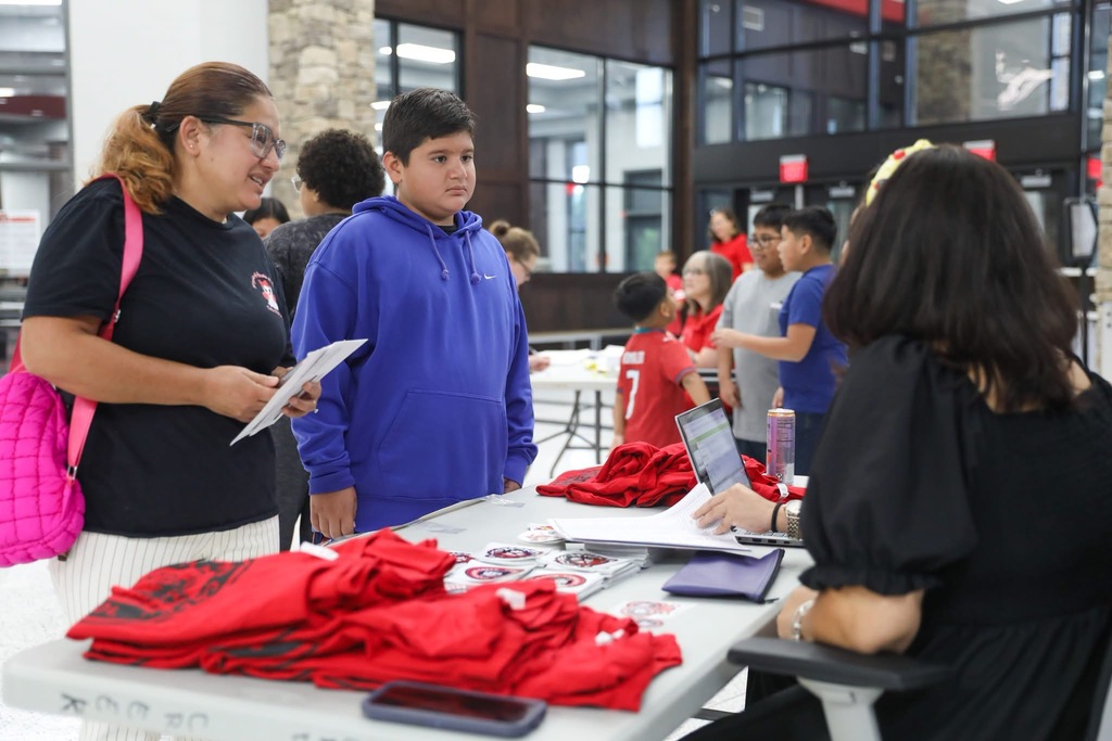 A woman wearing glasses and a boy in a blue hooded sweatshirt talking together in a bustling school atrium filled with students and tables.