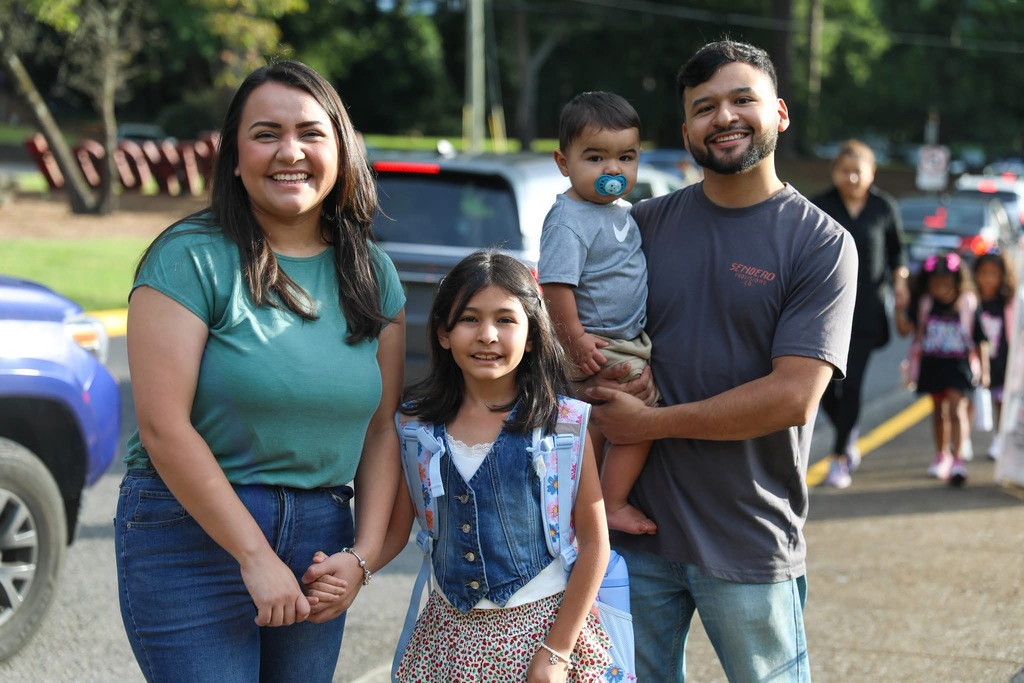 A smiling family posing together outdoors, with a woman in a teal shirt, a young girl, and a man holding a toddler with a pacifier.