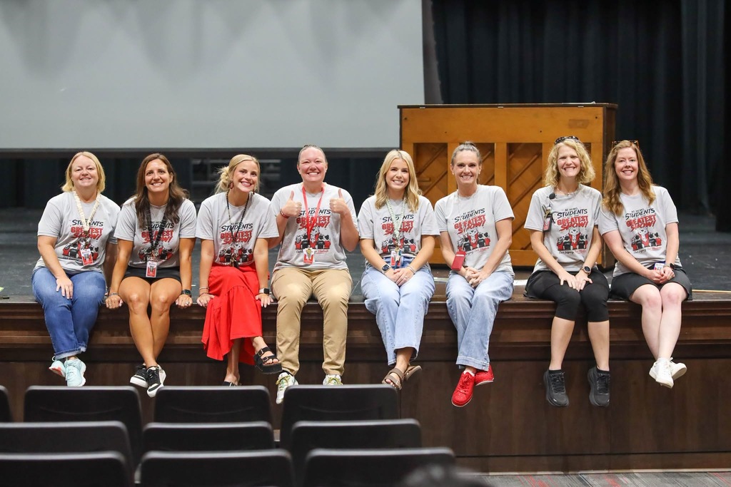 A group of eight smiling teachers and staff members wearing matching white spirit shirts seated in a row on a stage in front of a wooden podium and dark curtains.