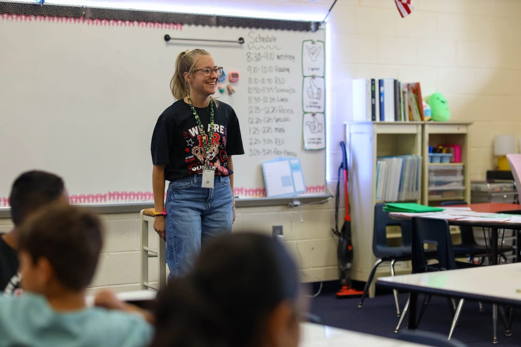 A smiling teacher with blonde hair and glasses stands in front of a classroom whiteboard and bookshelf.
