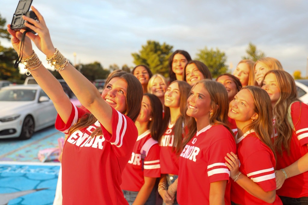 A group of smiling high school girls posing closely together outdoors for a selfie taken with a handheld digital camera.