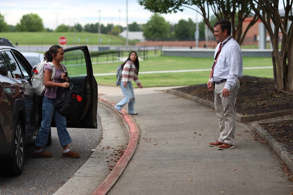 A school staff member in a white shirt and tie stands outside greeting students as they exit a car with their backpacks.