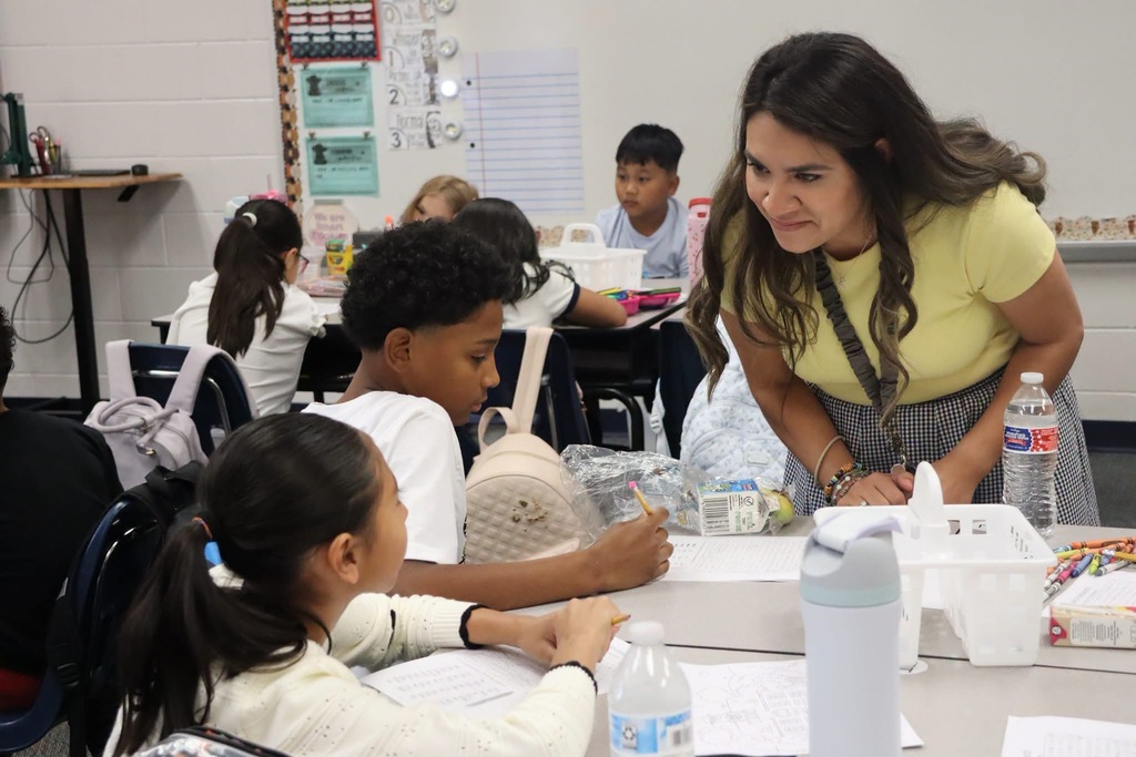 A teacher in a yellow shirt leans over a classroom table to assist two students who are writing and working together.