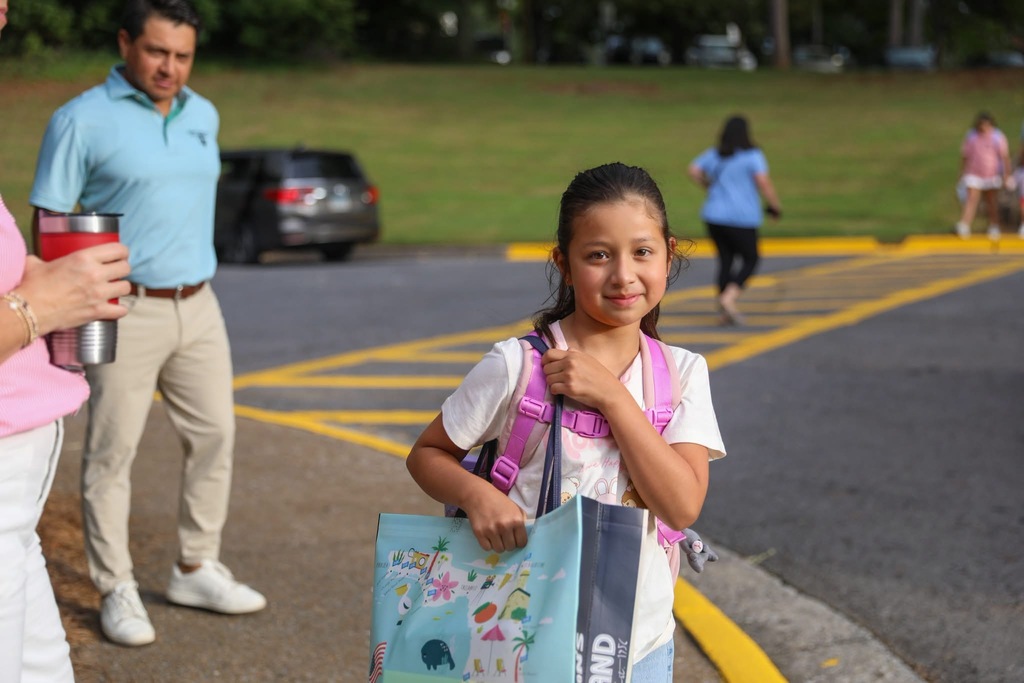 A young girl smiles at the camera as an adult carrying a drink cup walks past in an outdoor school parking lot or drop-off area