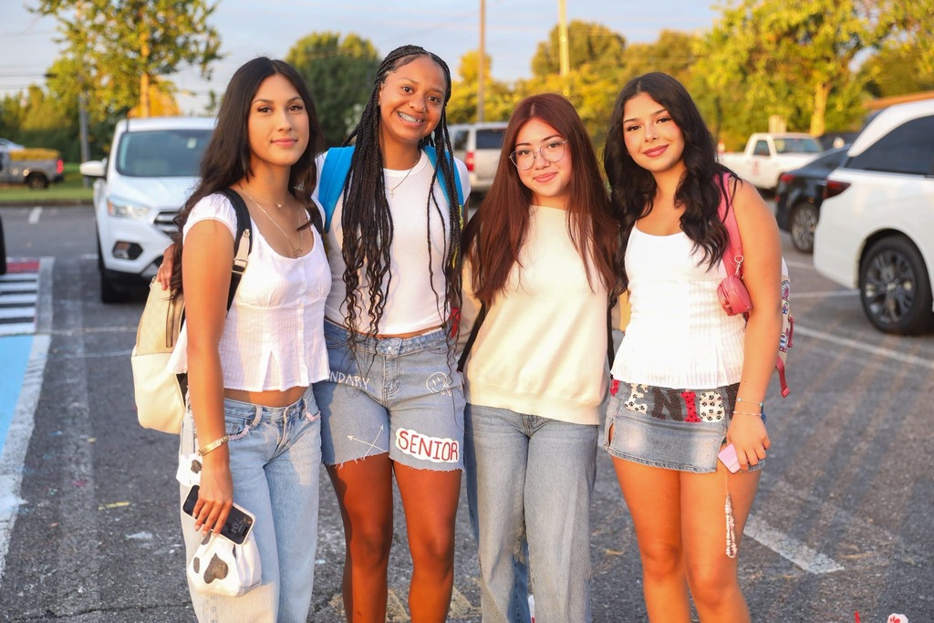 Four smiling young women posing together outdoors in front of a parking lot and trees.