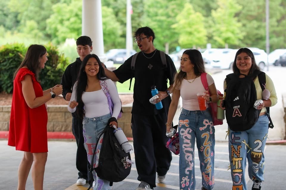 A group of six smiling high school students carrying backpacks walking and talking together outdoors in front of trees and a parking lot.