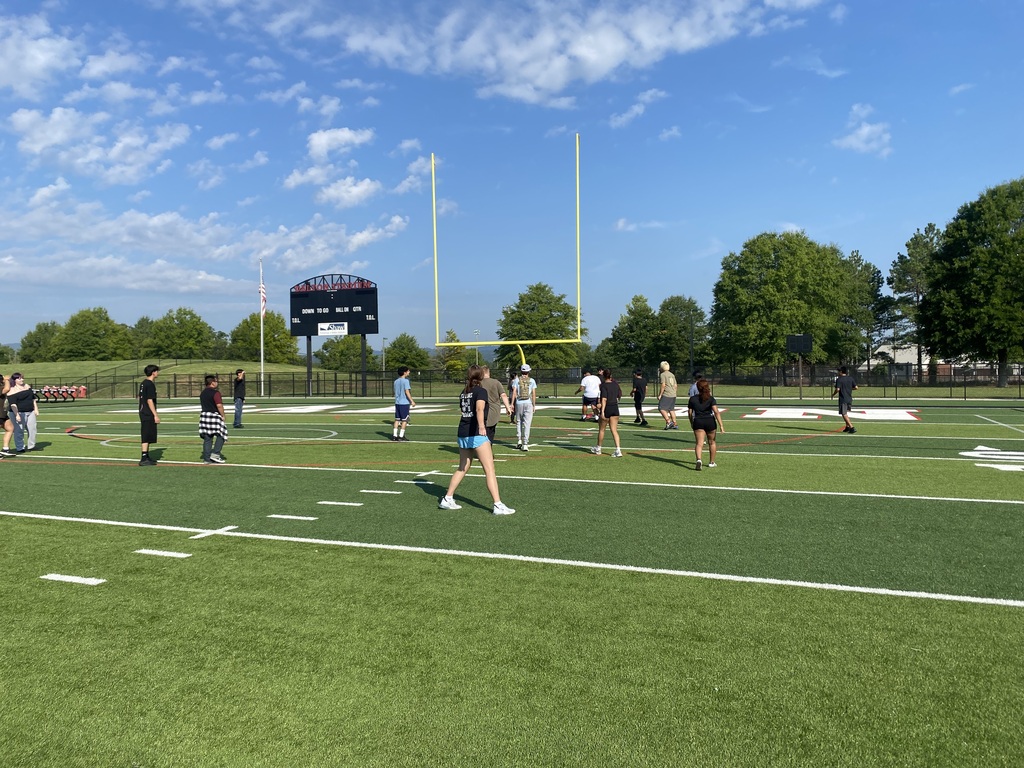 A wide view of a group of students scattered across a green outdoor football field on a sunny morning.