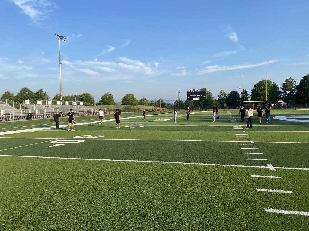 A wide view of a group of students scattered across a green outdoor football field on a sunny morning.