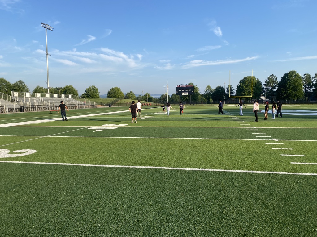 A wide view of a group of students scattered across a green outdoor football field on a sunny morning.