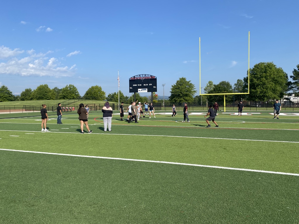 A wide view of a group of students scattered across a green outdoor football field on a sunny morning.