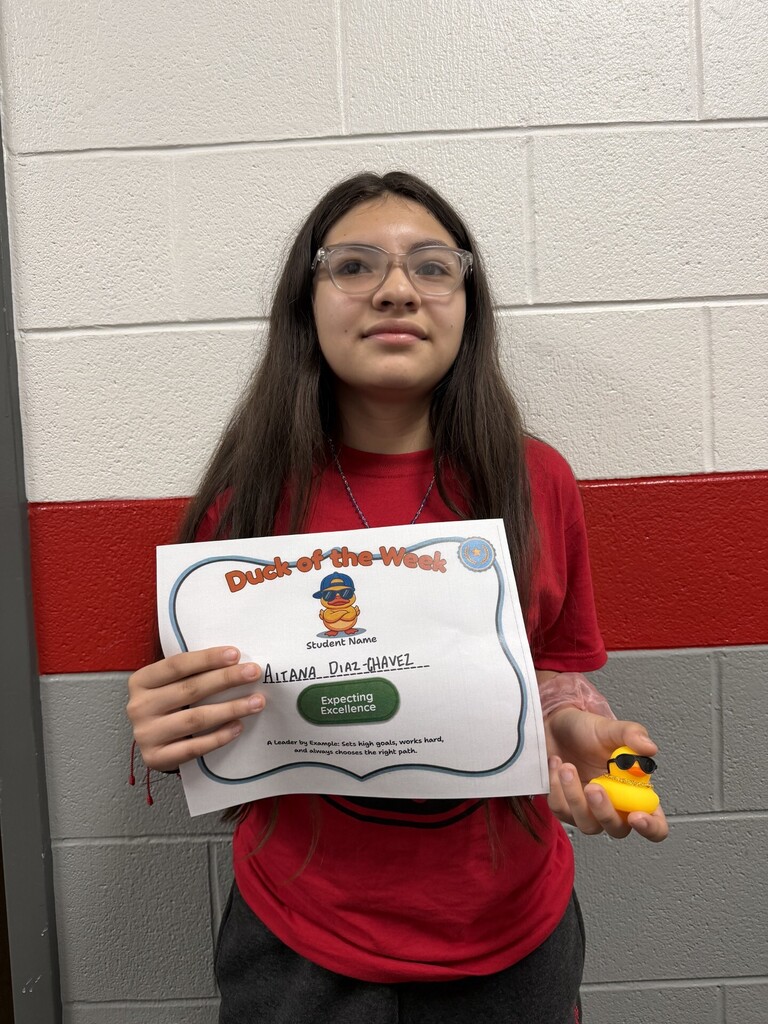 Student stands in front of a school hallway wall holding a "Duck of the Week" certificate and a small yellow duck award. The certificate recognizes Aitana Diaz-Chavez for "Expecting Excellence," with text describing a leader who sets high goals, works hard, and chooses the right path.