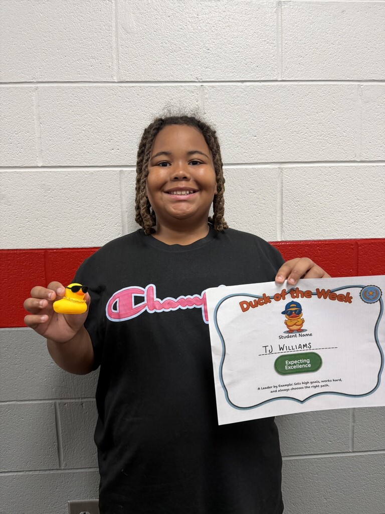 Student stands in front of a school hallway wall holding a "Duck of the Week" certificate and a small yellow duck award. The certificate recognizes TJ Williams for "Expecting Excellence," with text describing a leader who sets high goals, works hard, and chooses the right path.