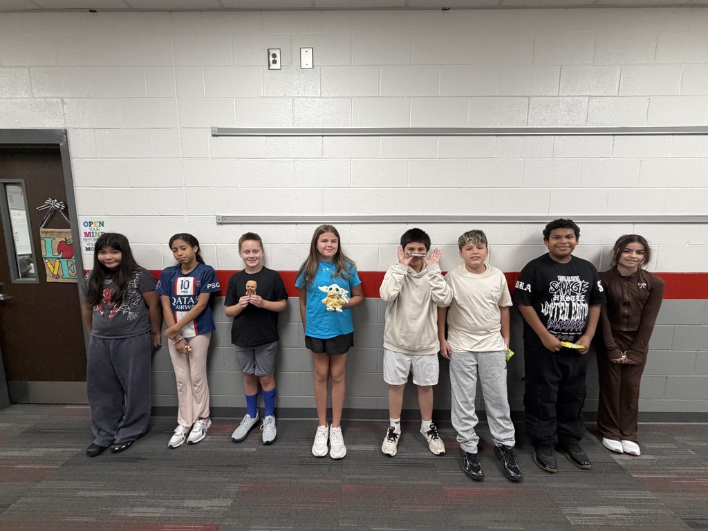 Group photo of eight students standing along a school hallway wall after being recognized as members of the Jedi team for the week. Several students are holding recognition items associated with the Jedi of the Week program. The students are lined up side by side in front of a gray and red wall in the school hallway.