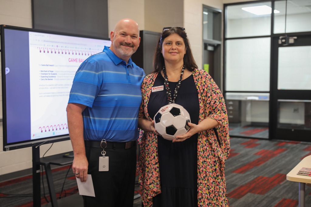 A bald man in a blue striped polo shirt stands beside a woman wearing a black dress and a colorful floral cardigan inside a school facility. The woman smiles as she holds a black-and-white soccer ball award. To their left, a large interactive display screen presents a "Game Ball" recognition page.