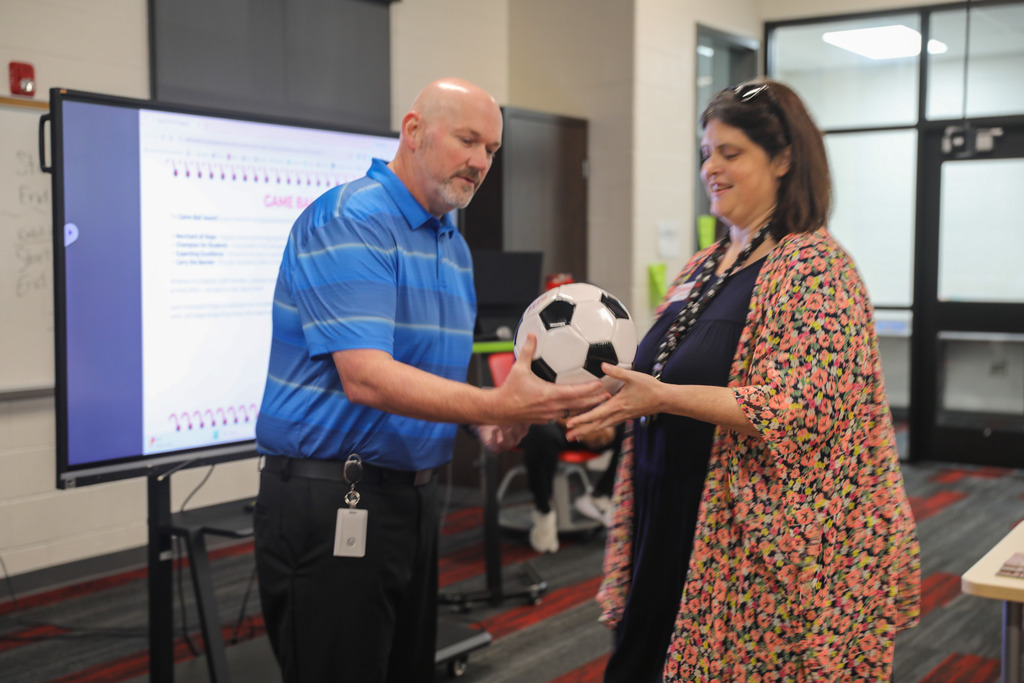 A bald man in a blue striped polo shirt presents a black-and-white soccer ball award to a woman wearing a dark dress and a colorful floral cardigan. They stand together in an indoor meeting space with an interactive digital display in the background showing a recognition program screen.