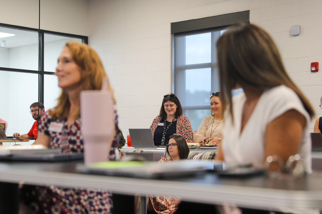 Participants seated at tables in a brightly lit room during a meeting or workshop. In the background focus, a woman in a floral cardigan works on a laptop next to another attendee, while colleagues sit in the foreground at desks equipped with computers and notebooks.