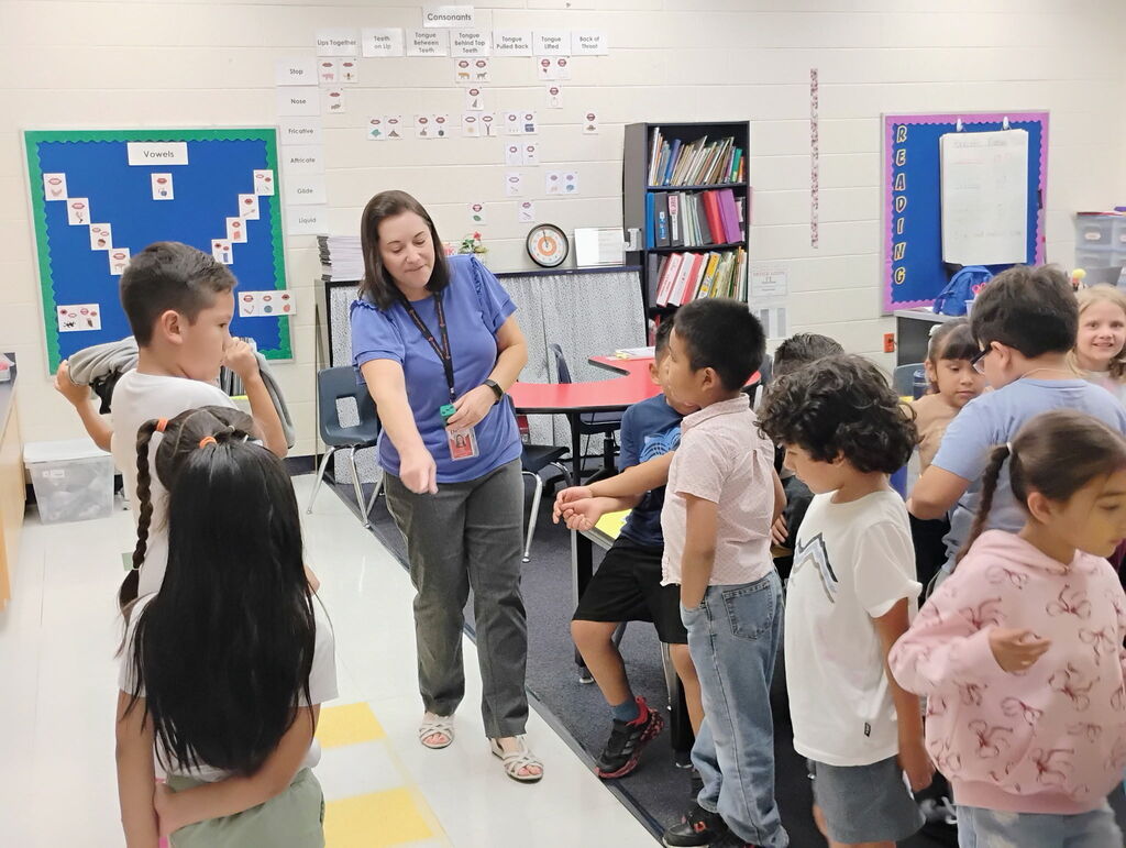 A female teacher in a blue top and lanyards stands over a group of elementary students participating in a structured line-up activity inside a classroom. To the left, colorful student backpacks hang on hooks along the wall near educational posters.