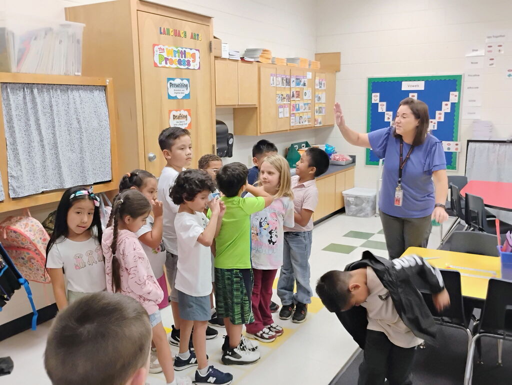 A female teacher in a blue top and lanyards stands over a group of elementary students participating in a structured line-up activity inside a classroom. To the left, colorful student backpacks hang on hooks along the wall near educational posters.
