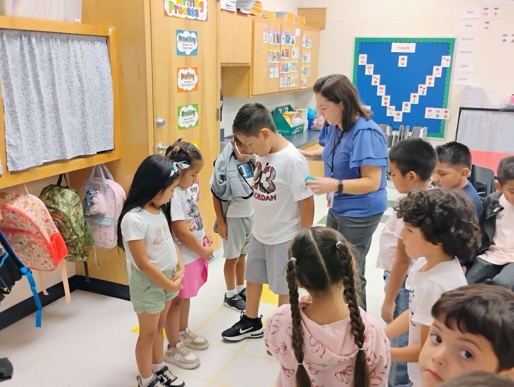 A female teacher in a blue top and lanyards stands over a group of elementary students participating in a structured line-up activity inside a classroom. To the left, colorful student backpacks hang on hooks along the wall near educational posters.