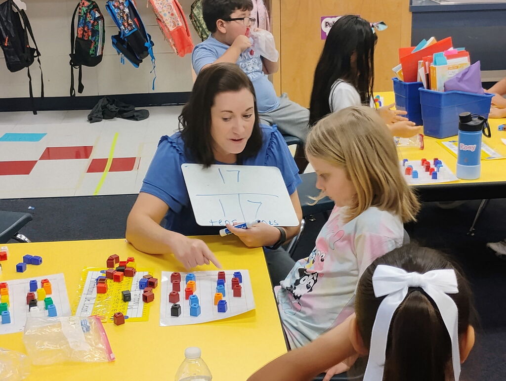 A teacher holds up a small handheld whiteboard showing the number 17 broken down into tens and ones, pointing to counting blocks on a worksheet in front of a young student seated at a yellow desk.