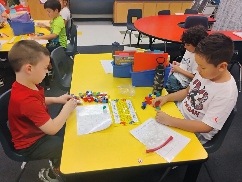 Two elementary school boys sit across from each other at a bright yellow table, organizing colorful plastic connecting blocks on math worksheets.