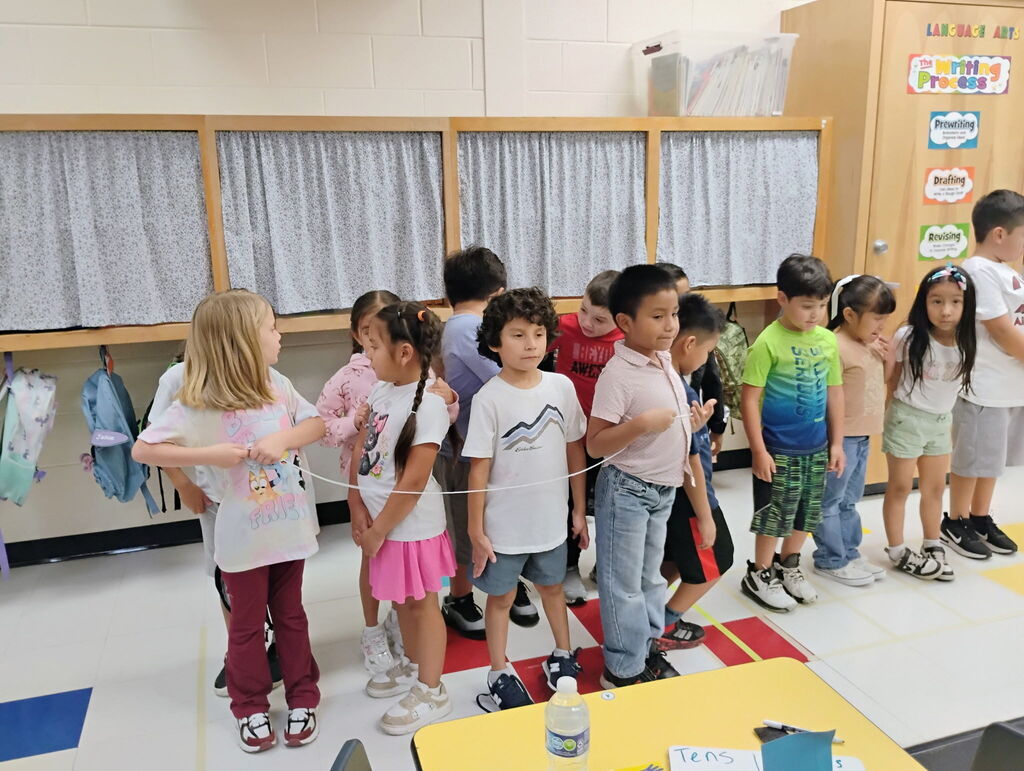 A female teacher in a blue top and lanyards stands over a group of elementary students participating in a structured line-up activity inside a classroom. To the left, colorful student backpacks hang on hooks along the wall near educational posters.