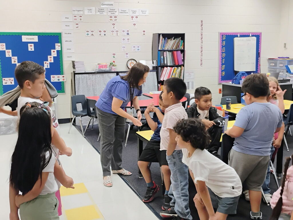 A female teacher in a blue top and lanyards stands over a group of elementary students participating in a structured line-up activity inside a classroom. To the left, colorful student backpacks hang on hooks along the wall near educational posters.