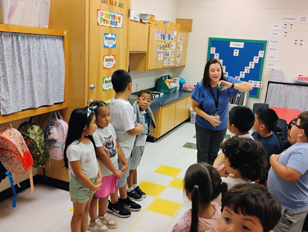 A female teacher in a blue top and lanyards stands over a group of elementary students participating in a structured line-up activity inside a classroom. To the left, colorful student backpacks hang on hooks along the wall near educational posters.