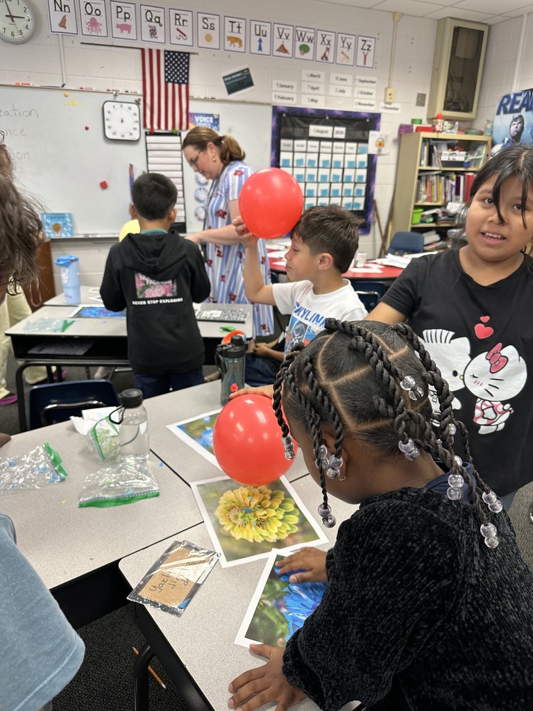 Students gather around desks in an elementary classroom during a science activity. In the foreground, a girl with braided hair examines a picture of a flower on her desk while holding a red balloon overhead, while another student in a white shirt rubs a red balloon against his head to generate static electricity.