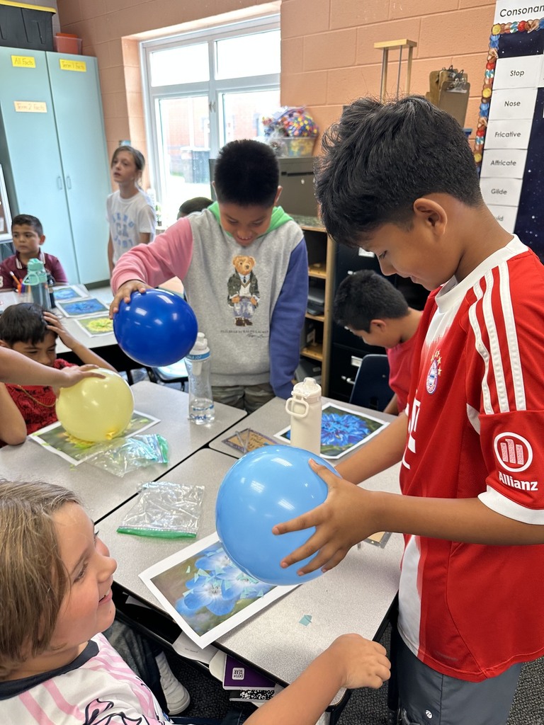 A group of elementary students work around classroom desks holding inflated blue and yellow balloons. One student in a red and white jersey holds a light blue balloon over a picture of a flower to attract tissue paper pieces using static electricity, while nearby peers observe and participate.