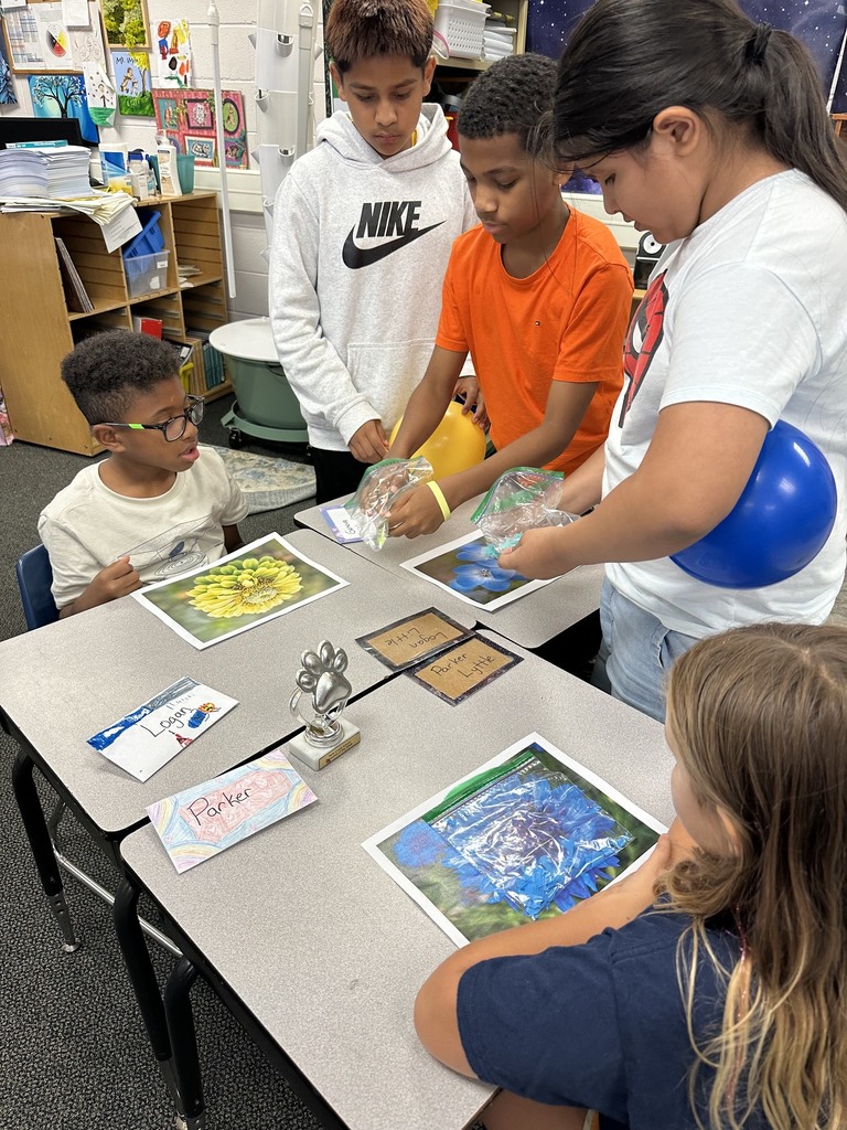 Four elementary students gather around classroom desks examining pictures of flowers and plastic bags. A student on the right holds a blue balloon, while two other students open plastic bags to test static electricity effects on colorful paper pieces scattered across flower images.