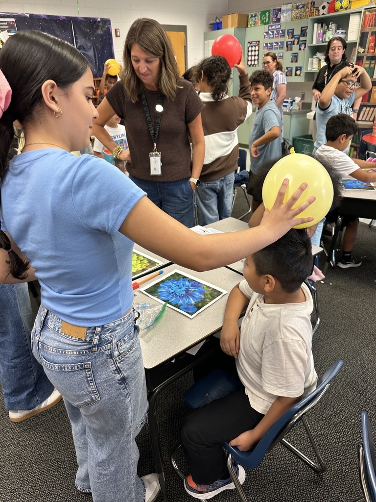 A girl wearing a blue shirt places a yellow inflated balloon onto a seated boy’s head during a science lesson on static electricity. Teachers and other students observe in the background as participants work at tables featuring flower prints and learning materials.