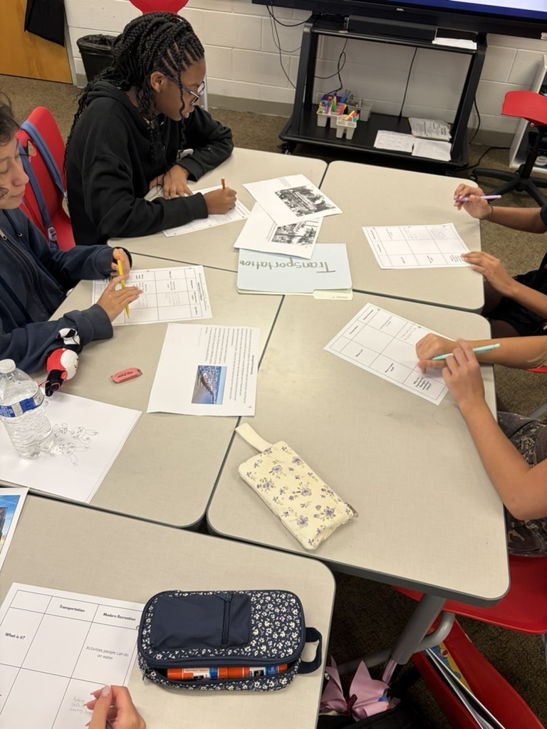 5 students sitting at desks in a cluster in a classroom working on an assignment