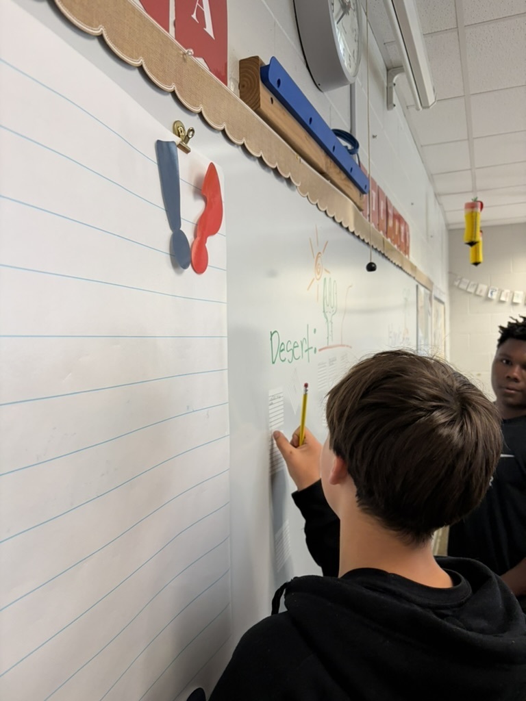 2 students wearing black shirts writing on a whiteboard in Georgia Studies class.