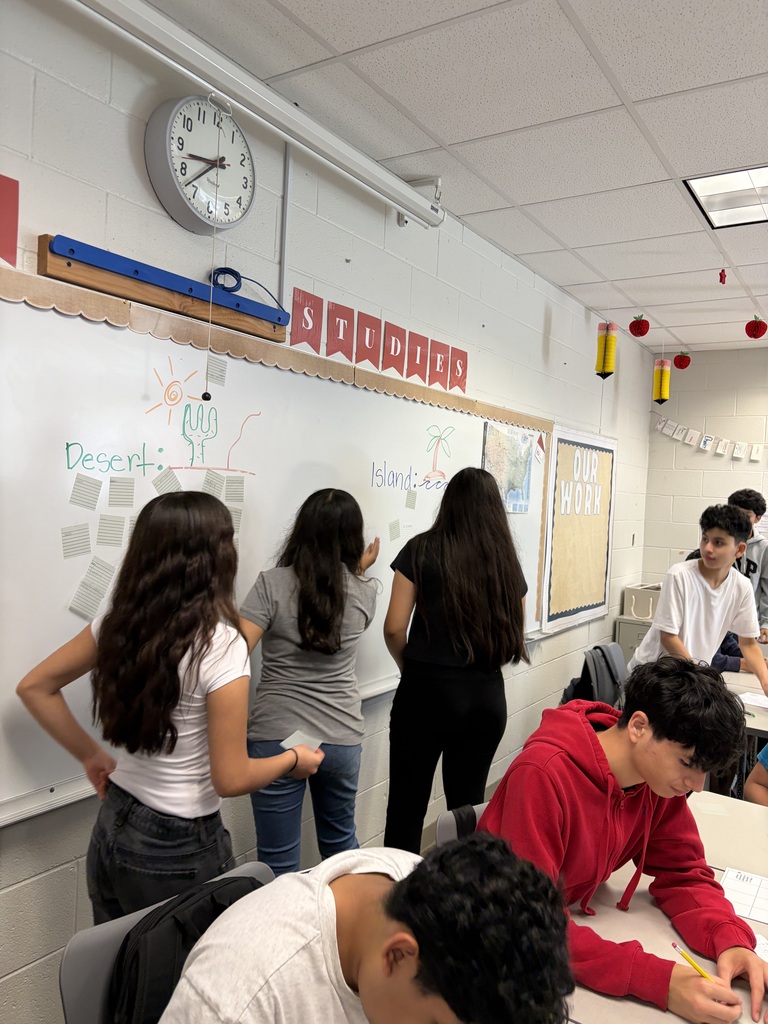 several students working in a classroom. 3 girls standing a the whiteboard while 2 boys look at what they are writing. 2 boys sitting at desks writing.