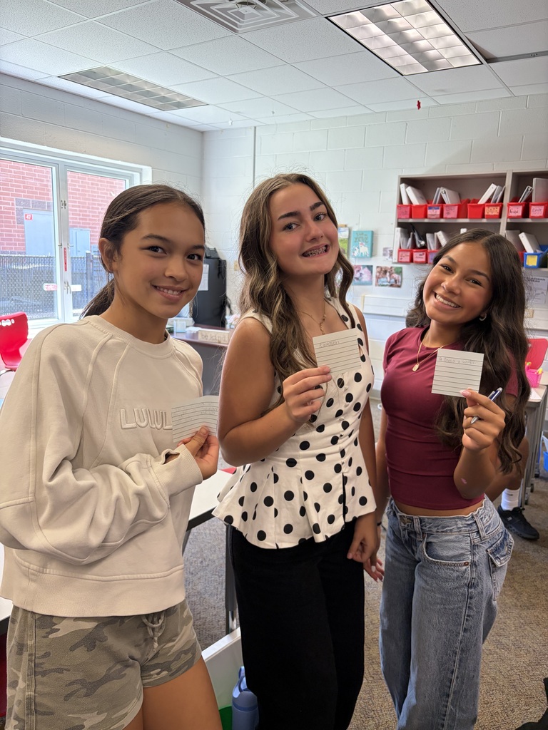3 girls standing in a classroom, each holding up a white card and smiling 