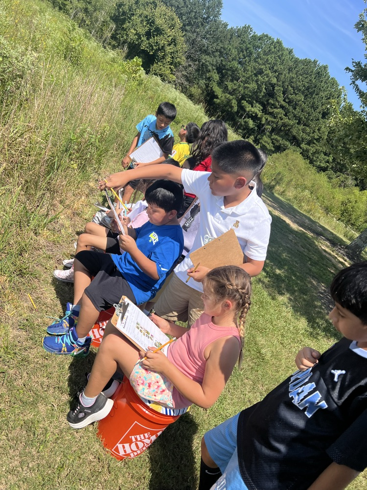 A group of students sitting on orange buckets and taking notes on clipboards in a grassy field.