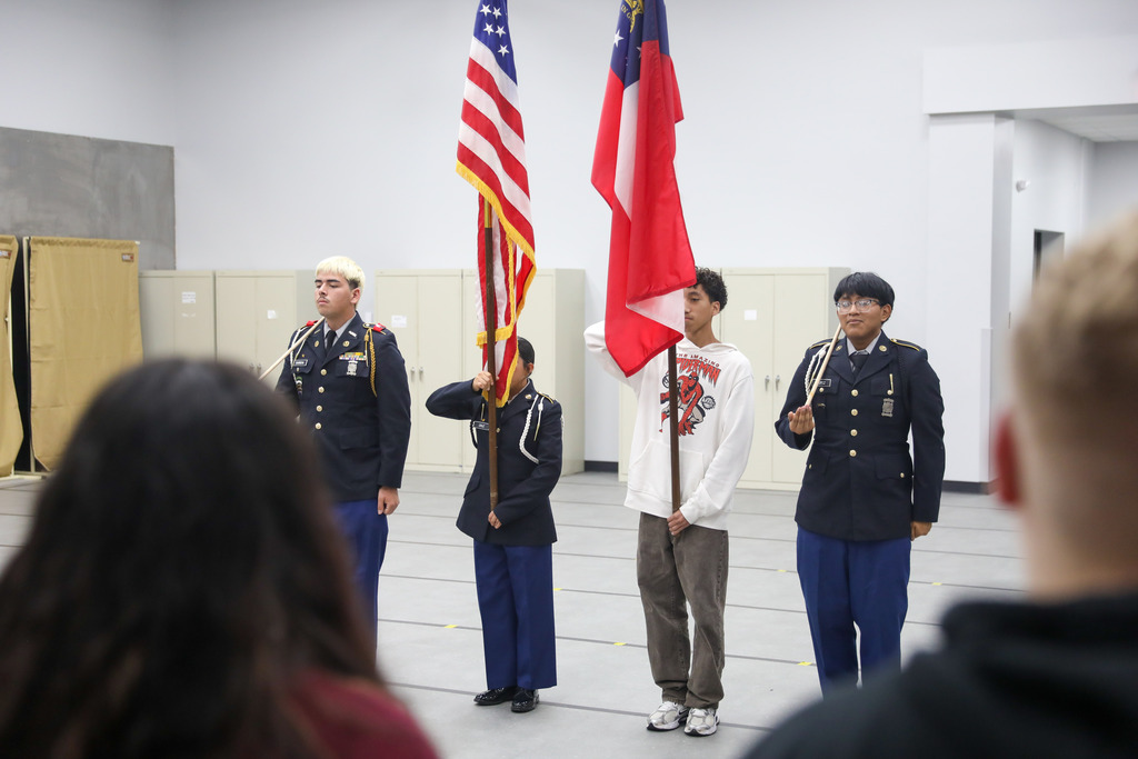 Four students stand in formation on a marked gray floor during color guard practice. Two students wear JROTC dress uniforms and hold ceremonial rifles, while a female cadet in uniform holds the U.S. flag and a male student in a white Spider-Man hoodie holds the Georgia state flag.