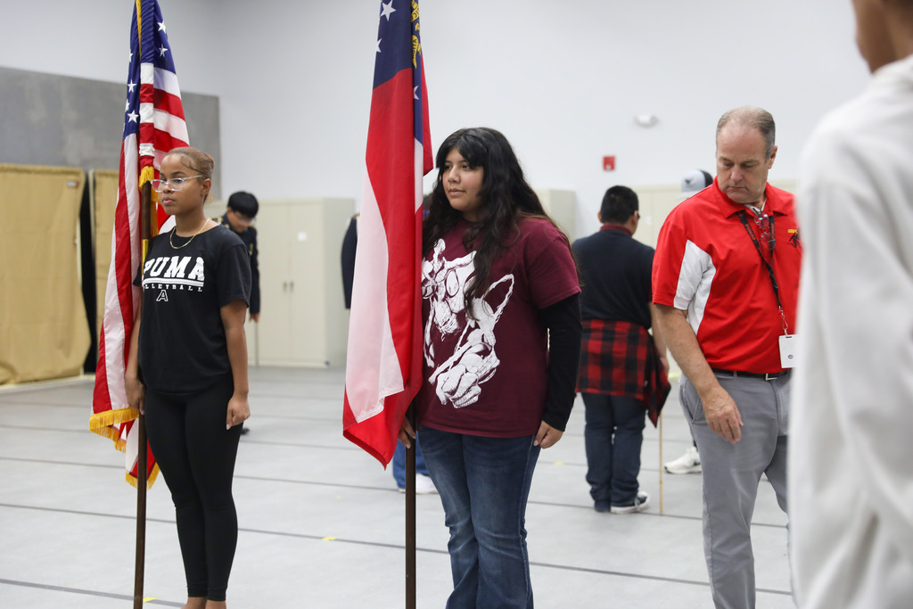 Two female students stand in formation on a marked gray floor holding flags during a practice session. The student on the left holds the United States flag, and the student on the right holds the Georgia state flag, while an adult instructor in a red polo shirt observes nearby.