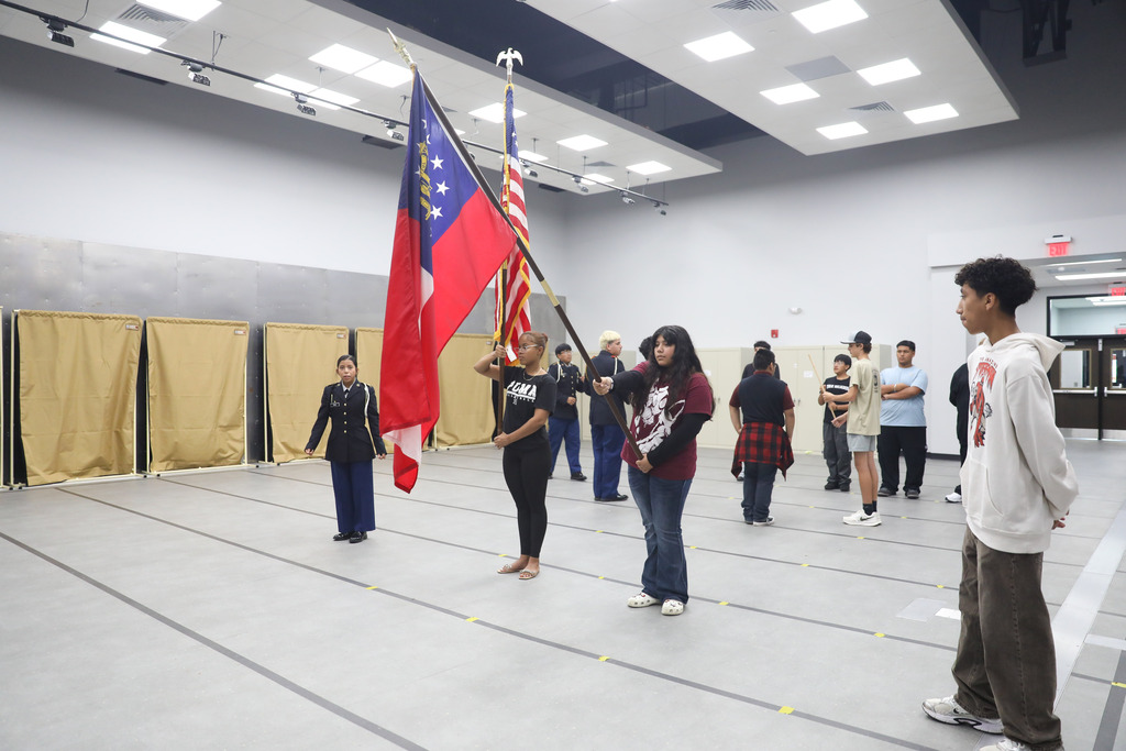 Multiple students are spread across a large practice room with a taped gray floor. In the center, two female students practice holding the U.S. and Georgia state flags aloft, while other students in the background practice with wooden ceremonial rifles under the supervision of an instructor.