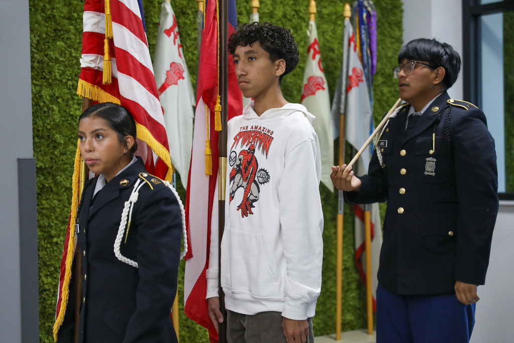 Three students practice color guard drill in a school hallway with a green moss wall in the background. A female JROTC cadet holds the U.S. flag, a male student in a white hoodie holds the Georgia flag, and a male cadet in uniform stands behind them holding a ceremonial rifle.
