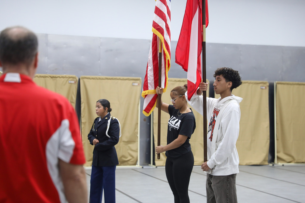 A female student in a black shirt and a male student in a white hoodie hold the United States and Georgia state flags at attention. An instructor in a red polo shirt stands in the foreground observing their technique during a color guard practice session.