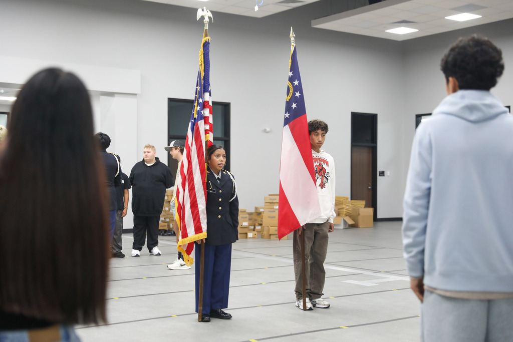 A female JROTC cadet in uniform holds the United States flag while a male student in a white hoodie holds the Georgia state flag. They stand at attention on a taped gray floor in a large, bright room while other students watch.