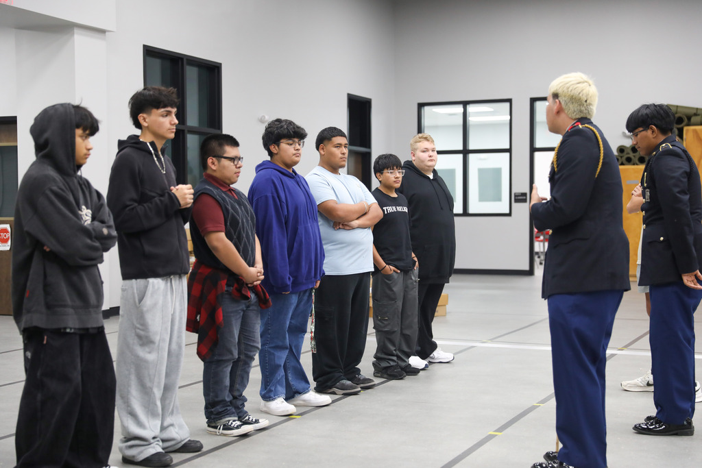 A JROTC cadet in full dress uniform stands addressing a line of six male high school students in casual clothing. They are standing on a gray floor marked with black tape lines inside a large, brightly lit multi-purpose room.