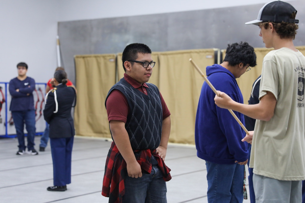 Several students in casual clothing practice handling wooden ceremonial drill rifles in a large room with a gray floor. In the foreground, a student in a red polo shirt and argyle sweater vest watches while others practice movements behind him.
