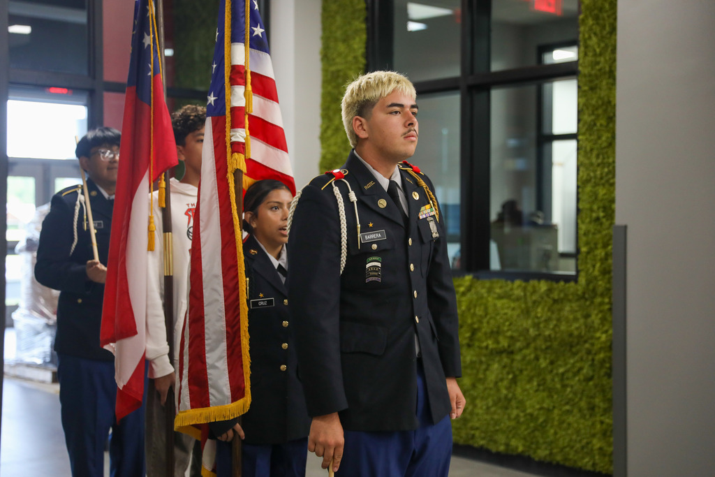 A JROTC cadet in a dark blue dress uniform with a yellow shoulder cord stands at attention in the foreground. Behind him, a female cadet holds the United States flag, while another cadet holds the Georgia state flag during a color guard practice in a hallway featuring a moss green accent wall.